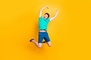 Fototapeta na wymiar Full size length body picture of handsome curly-haired playful young guy wearing casual green t-shirt, shorts, shoes, jumping, hands up, celebrating prize winning. Isolated over yellow background
