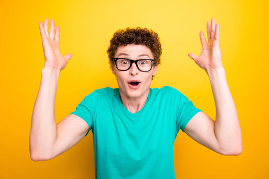 Handsome curly-haired shocked young guy wearing casual green t-shirt and glasses, showing surprised gesture. Isolated over yellow background