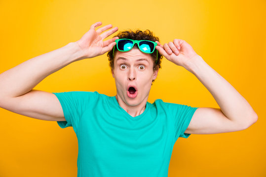 Young attractive handsome shocked guy wearing casual green polo t-shirt and sun glasses, raising glasses up showing wow facial expression. Isolated over vivid shine bright yellow background