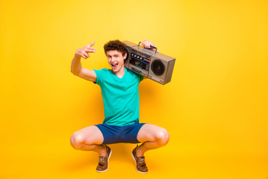 Full height body size handsome curly-haired funny young guy carrying boombox, wearing casual green t-shirt, shorts, shoes, singing. Isolated over yellow background