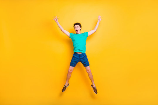 Full size length body picture of handsome curly-haired funny young guy wearing casual green t-shirt, shorts, shoes, jumping in air, hands up, star figure. Isolated over yellow background