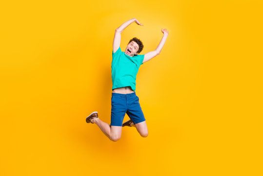 Full size length body picture of handsome curly-haired playful young guy wearing casual green t-shirt, shorts, shoes, jumping, hands up, celebrating prize winning. Isolated over yellow background