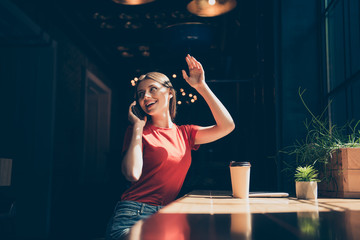 Attractive nice smiling young girl wearing casual sitting in cafeteria, talking on phone, waving, waiting for friend, drinking coffee, dark interior