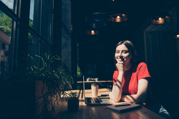 Attractive nice smiling young girl freelancer student wearing casual sitting in cafeteria, working remotely, writing notes in pad, drinking coffee, dark interior