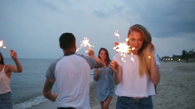 Friends walking, dancing and having fun during night party at the seaside with bengal sparkler lights in their hands. Young teenagers partying on the beach with fireworks. Slow motion steadycam shot.