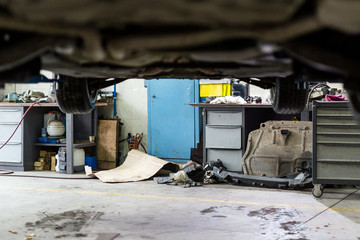 Car hanged on lift at service station. Dirty vehicle  maintenance center. Disorder, mud, stains and chaos. Unprofessional clumsy automobile workshop - obrazy, fototapety, plakaty