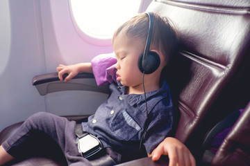Fototapeta premium Cute little Asian 24 months / 2 years old toddler baby boy child sleeping on Airplane, Toddler boy sitting with safety belt on wearing headphones while traveling in airplane, Kids Fly Safe concept