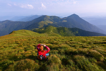 Among the nice green hummocks there is a red hair girl sitting at halt and watching the horizon with the beatufil mountains. Sky with interesting clouds. Summer evening full of dreams.