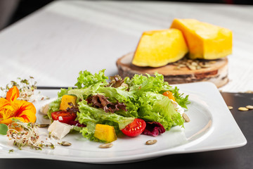 European Italian salad of lettuce, cherry tomatoes, pumpkin, edible pumpkin flowers, and pumpkin seeds, in a white plate. Copy space, selective focus