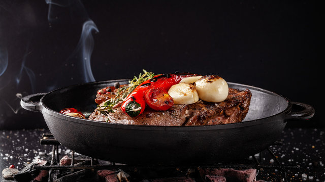 concept of the process of cooking meat. The steak is roasted on charcoal in a cast-iron frying pan, with spices, garlic, hot red pepper, squarish, thyme. image background. Copy space, selective focus