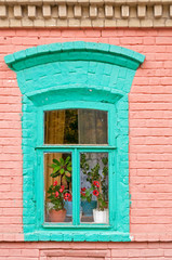 Green arched window in pink brick wall of old building and red blossoming flowers in flowerpots on windowsill behind glass. Gorodets, Nizhegorodsky region, Russia. 
