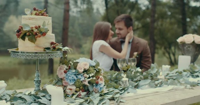 Close-up view at the tasty cake, bouquet, candles and leaves at table at the blurred background of the loving beautiful couple enjoying each other. Woman is stroking the face of man. Misty forest.