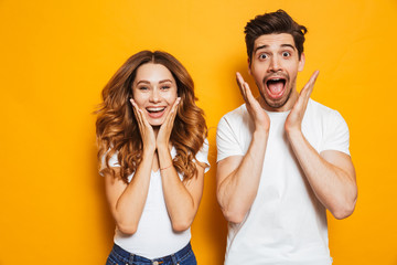 Photo of amazed couple man and woman in basic clothing screaming in surprise or delight and touching cheeks, isolated over yellow background