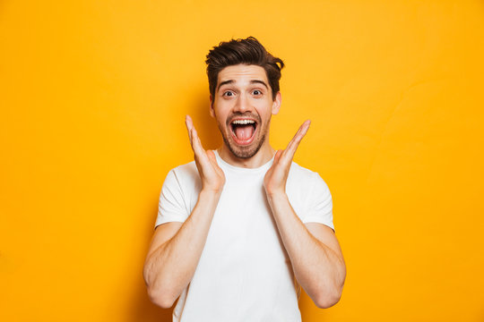 Photo of amazed man in basic clothing screaming in surprise or delight and raising arms, isolated over yellow background