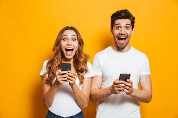 Photo of caucasian beautiful people man and woman screaming, while both using smartphones, isolated over yellow background