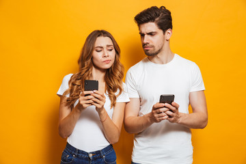 Image of curious man and woman frowning and peeking at each others cell phones, isolated over yellow background