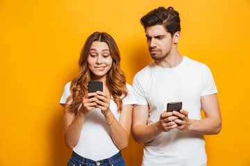 Image of curious man frowning and peeking at smartphone of his girlfriend, isolated over yellow background