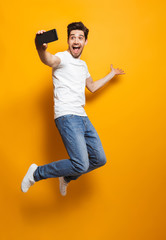Full length portrait of happy man with brown hair jumping and screaming while taking selfie on...