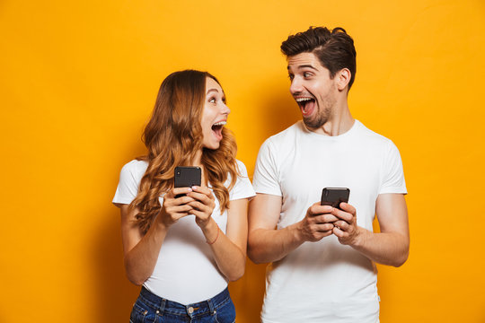 Photo of positive excited people man and woman screaming and looking at each other while both using mobile phones, isolated over yellow background