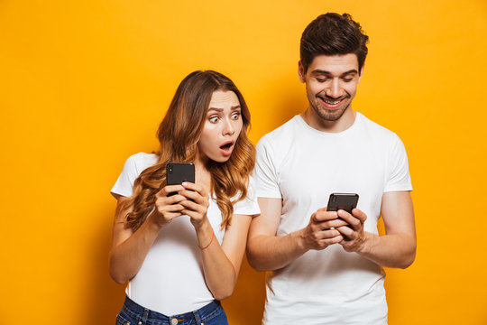 Image Of Young Woman Spying And Peeking At Smartphone Of Her Boyfriend, Isolated Over Yellow Background