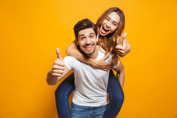 Image of joyous couple showing thumbs up while man piggybacking joyful woman, isolated over yellow...