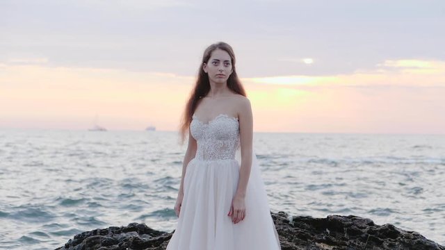Girl in wedding luxury dress posing on sea shore. Bride on a rocks. Beautiful waves near to her. Woman enjoying happy moments with nature. Sunrise or sunset time. Slow motion.