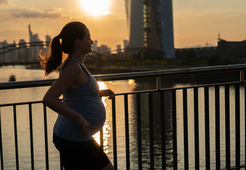Fototapeta premium Active pregnant woman running across the bridge with a beautiful view of the city of Frankfurt in Germany