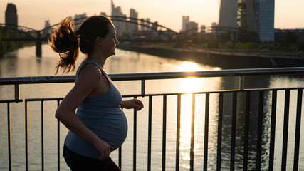 Active pregnant woman running across the bridge with a beautiful view of the city of Frankfurt in Germany - 215805357