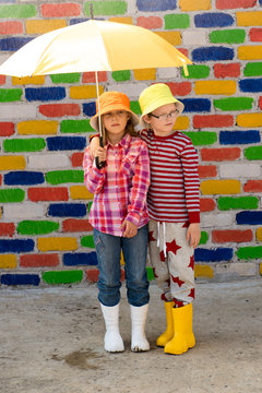 Happy children in colorful hats are sheltered from the rain under multicolored umbrellas in front of mottled brick wall. Fun during the summer holidays. Boys and girl have funny time. Copy space