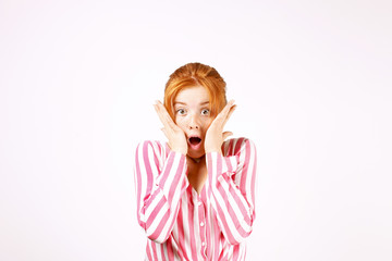 Beautiful redhead young woman looking scared, eyes wide open, hands on face. Pretty female with childish frightened facial expression, reaction to scary movie. White background, copy space, close up.