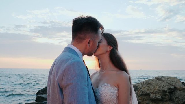 Beautiful young wedding couple standing on sea shore with rocks. Newlyweds spend time together: embrace, kiss and care for each other. Love concept.