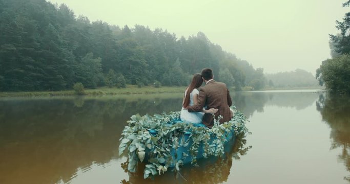 Rear view of the stylish couple in vintage sloth floating on romantic boat decorated with green herbs along the foggy river. Woman is putting her head on her lover shoulder. 4k.