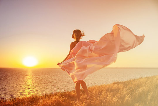 A girl with a pareo developing in the wind, standing on the beach, meets the sunrise and basks under the warm morning rays.
