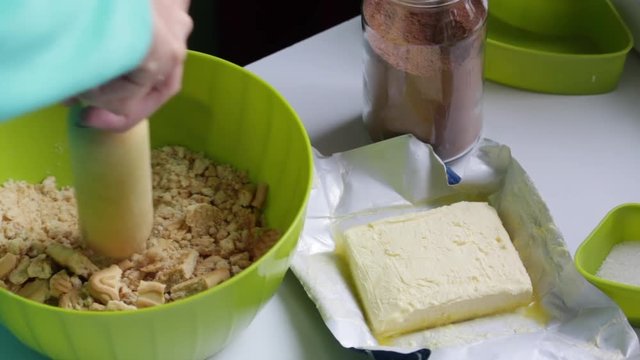 A woman shreds a biscuit with a rolling pin. Preparation of chocolate sweets from cocoa and biscuits. On the table is a ingredients. Biscuits, cocoa, butter and sugar in containers.