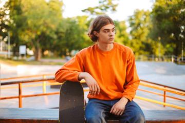 Young skater in orange pullover thoughtfully looking aside while leaning on skateboard with modern skatepark on background