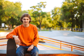 Young smiling skater in orange pullover joyfully looking in camera while leaning on skateboard with modern skatepark on background