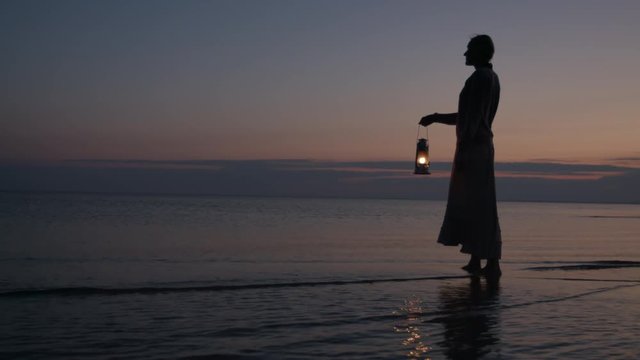 Girl in dress holding old kerosene lantern in her hand at beach after sunset at night