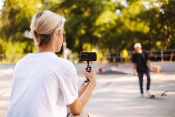 Pretty girl recording new video of young skater for vlog while spending time at skatepark