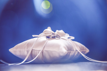 White Bridal Pillow and White Gold Wedding Rings with Light Flare on Dark Leather Sofa.