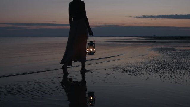 Girl in dress walking on beach and holding old kerosene lantern in her hand after sunset at night