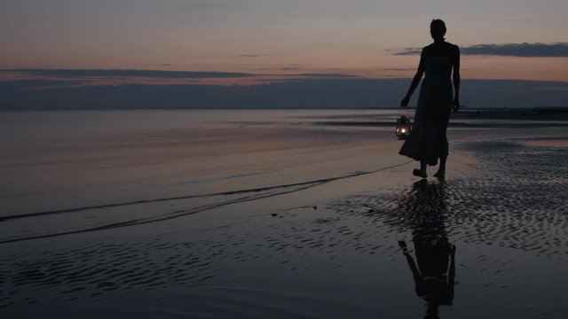 Girl in dress walking on beach and holding old kerosene lantern in her hand after sunset at night
