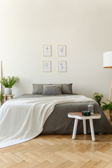 White blanket on grey bed in bright bedroom interior with posters and wooden table. Real photo