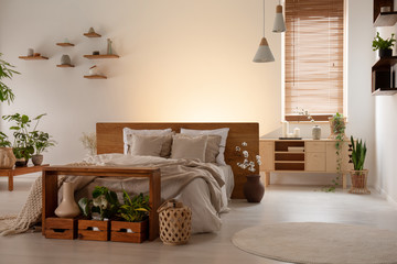 Simple double bed, table, boxes with plants, window blinds next to an empty wall in a bedroom...