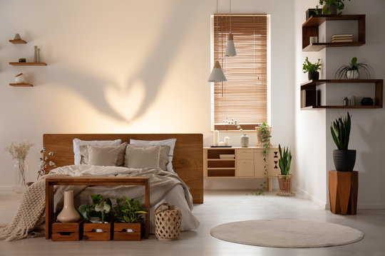 Real photo of a warm bedroom interior with a bed, table, wooden boxes, shadow on the wall and plants