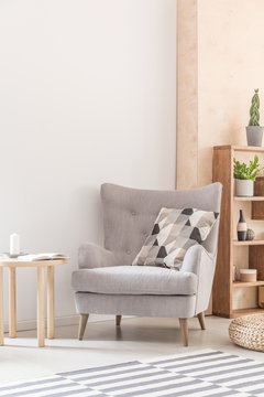 Comfy armchair with patterned pillow next to a wooden coffee table in a living room interior. Real photo. Place your poster