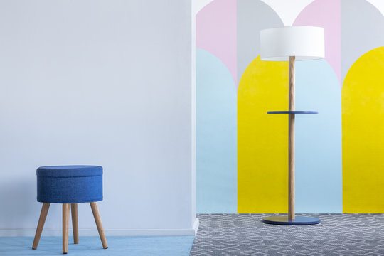 Real photo of a blue stool and a modern lamp in bright living room interior with colorful arches on the white wall. Place for your poster