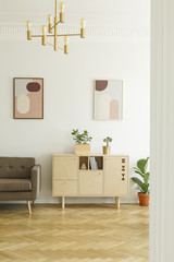 Posters on a white wall, comfy sofa and a simple, wooden cabinet in a real retro living room interior with herringbone parquet floor