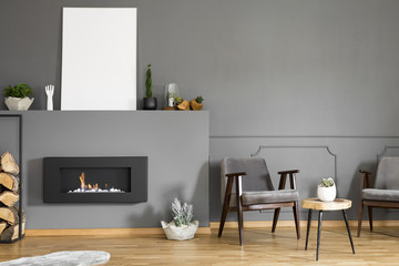 Real photo of two armchairs standing next to a stool and a fireplace with a mockup poster in dark living room interior