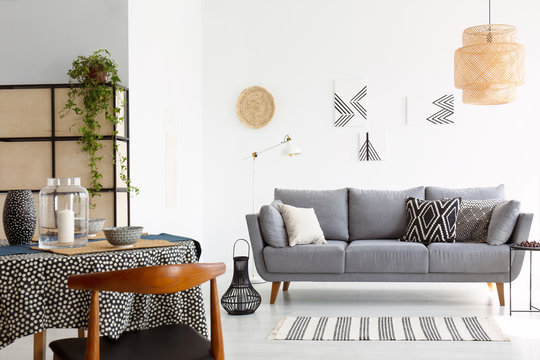 Real photo of a bright and cozy living room interior with patterned pillows on gray sofa and a wooden chair at a dining table