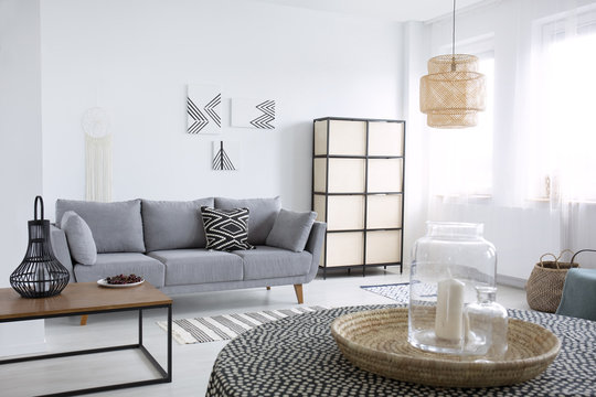 Real photo of a natural tray with glass vases and a candle on table with spotted tablecloth in the foreground of a scandi living room interior with gray sofa